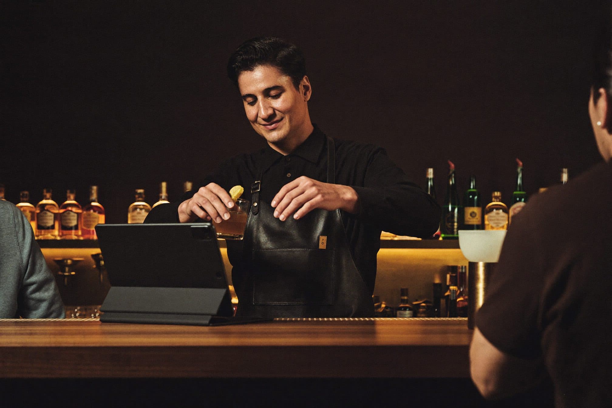 Un bartender termina un cóctel en la barra, con una tablet apoyada al lado.
