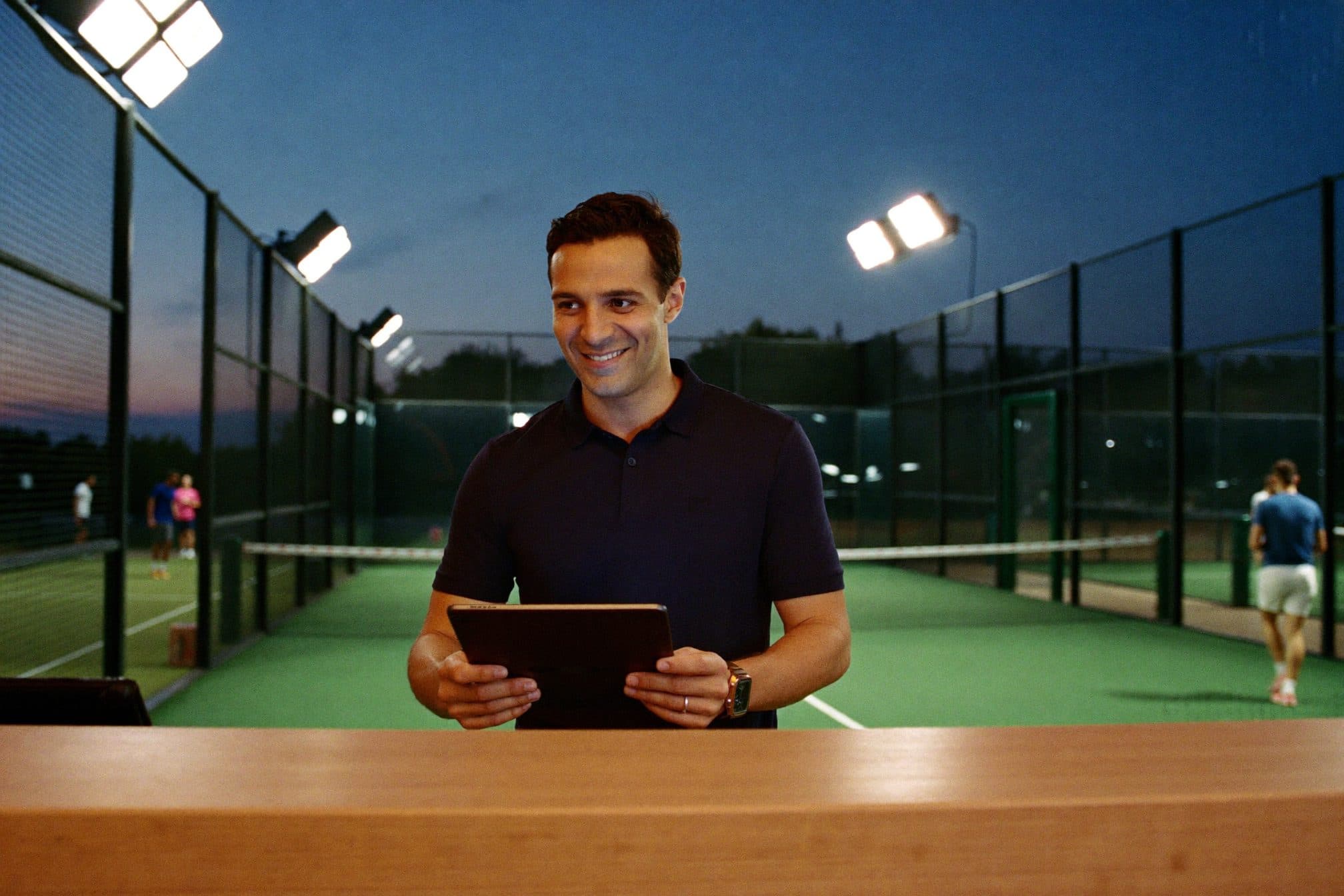 El encargado de un complejo de pádel revisa los turnos de la noche en una tablet, con las canchas iluminadas detrás.