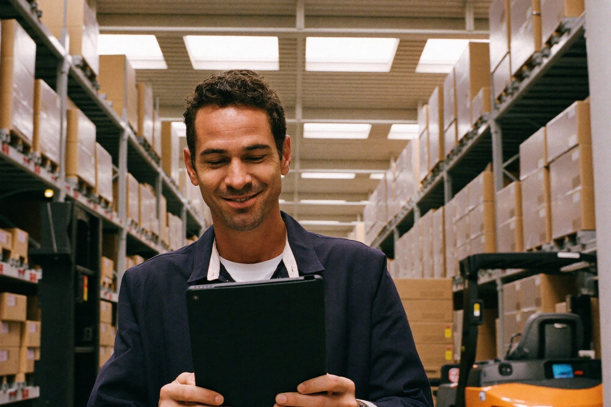 El encargado de un depósito revisa un pedido en una tablet, entre estanterías ordenadas.