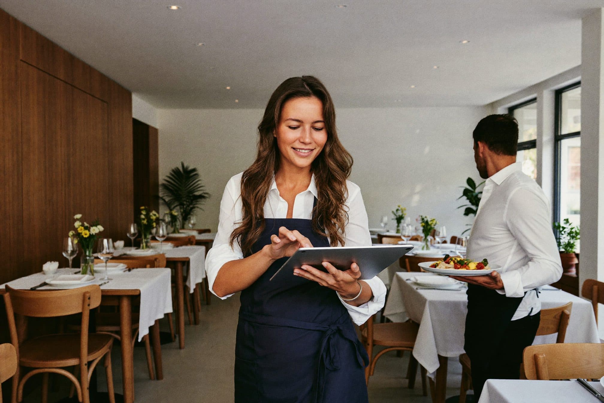La dueña de un restaurante revisa un pedido en una tablet, entre mesas con manteles blancos.